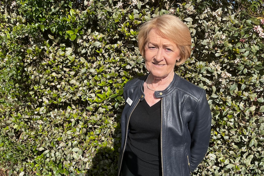 A older caucasian woman with short blonde hair and a black t-shirt and leather jacket stands in front of a hedge.