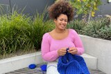 Yasmin sits outside and looks to the camera smiling while working on an electric blue cable jumper.