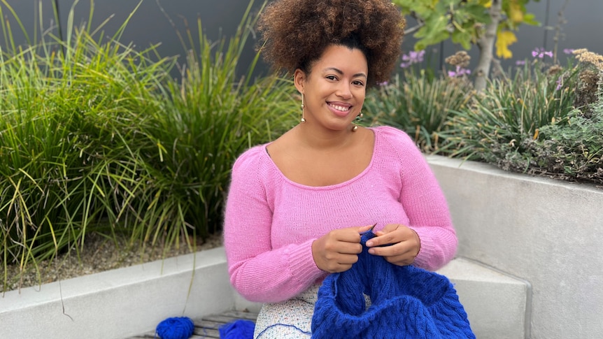 Yasmin sits outside and looks to the camera smiling while working on an electric blue cable jumper.