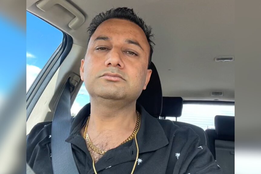A man with a black shirt and gold chains sits in a car