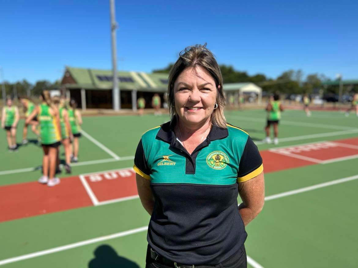 A woman standing on a netball court with a club shirt on.