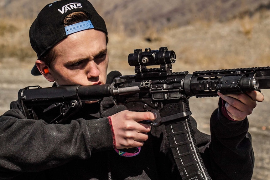 Image of a teenage boy with his hat turned backwards firing an AR-15 gun in an open space