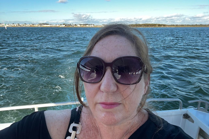 A middle-aged woman, dark blonde hair tied, few strands loose, wears large sunglasses, black tee, on boat in sea.