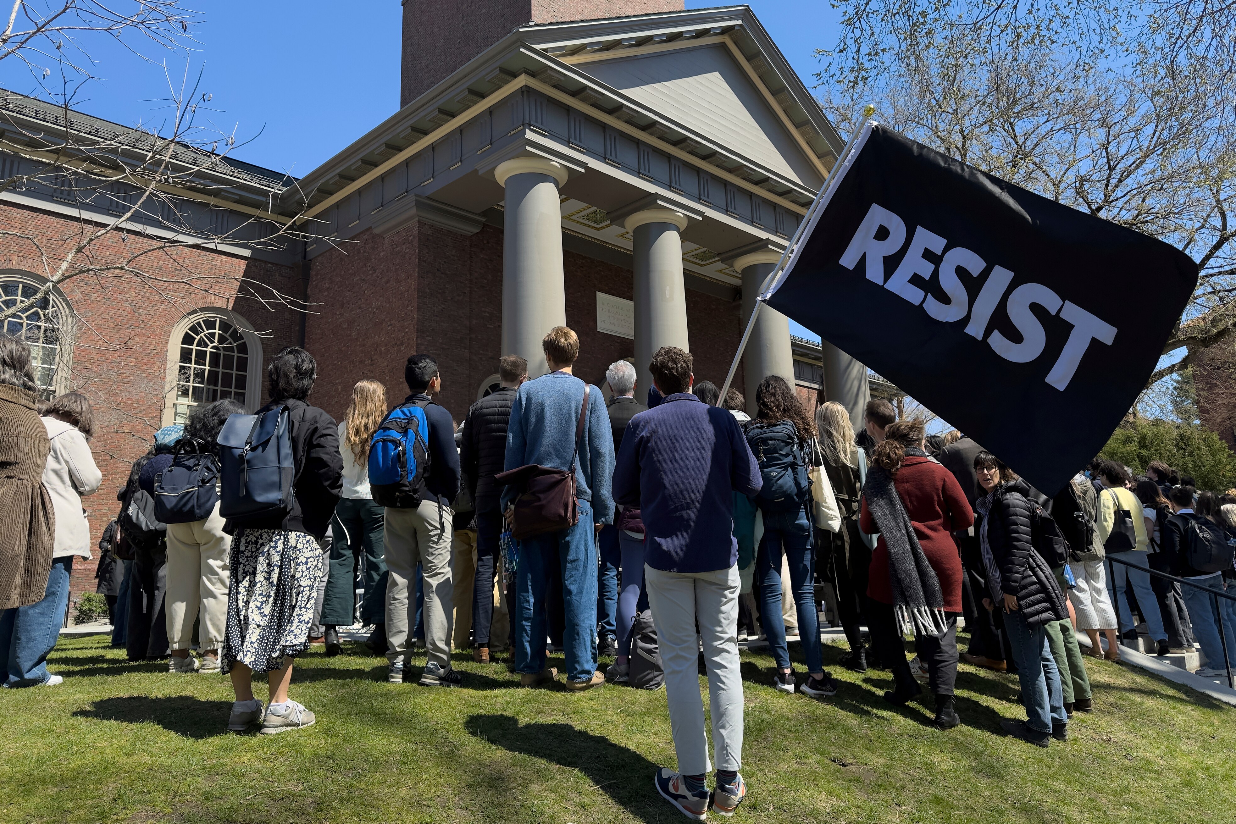 protesters stand outside Harvard building, one is holding a sign that says 'resist'