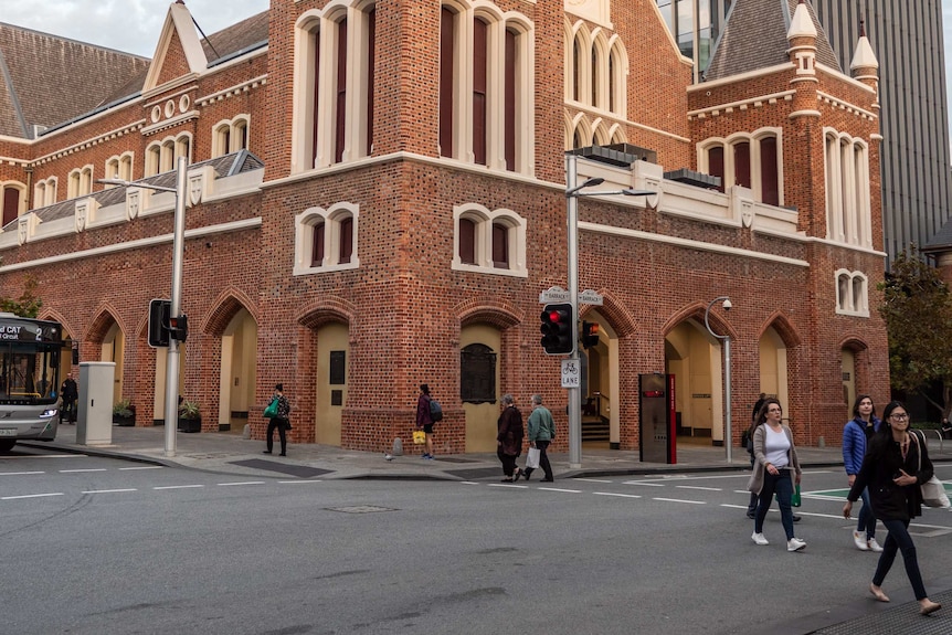 An intersection with pedestrians and cars, a Victorian brick building with gothic arches stands in the background.