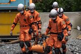 Five people in orange emergency gear and white helmets carry a stretcher with a dummy over rough terrain