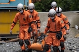 Five people in orange emergency gear and white helmets carry a stretcher with a dummy over rough terrain