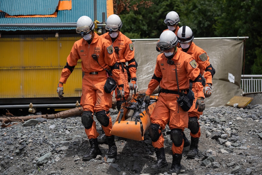 Five people in orange emergency gear and white helmets carry a stretcher with a dummy over rough terrain