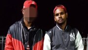 Two men wearing bomber jackets and red baseball caps backwards stand together at night.