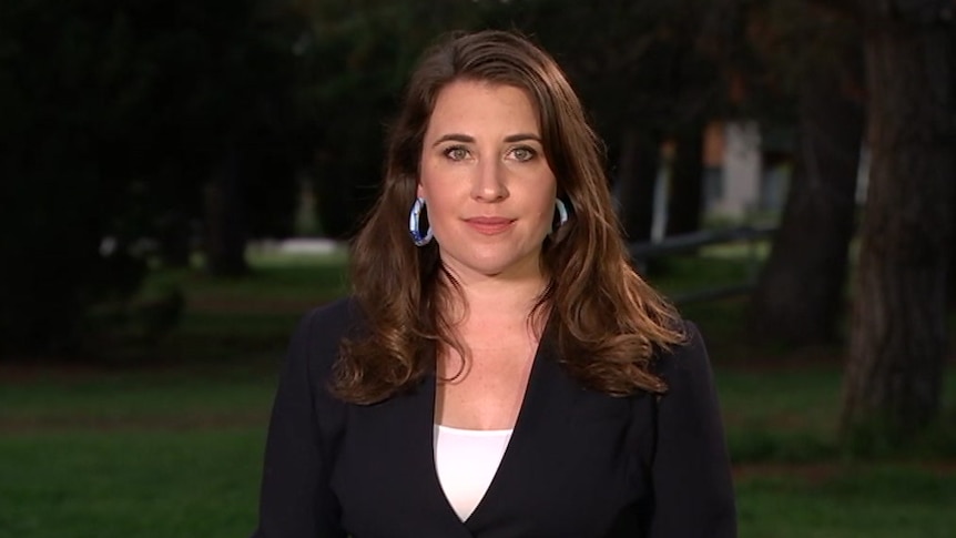 Woman with long brown hair wearing a black blazer and white shirt standing in front of a row of trees in the early evening.