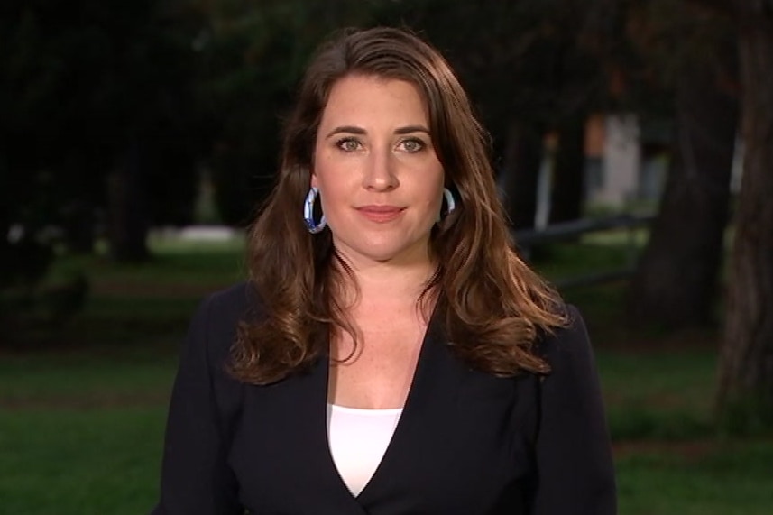 Woman with long brown hair wearing a black blazer and white shirt standing in front of a row of trees in the early evening.