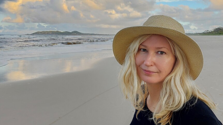 A woman in a black had with blonde hair stands on an empty beach.