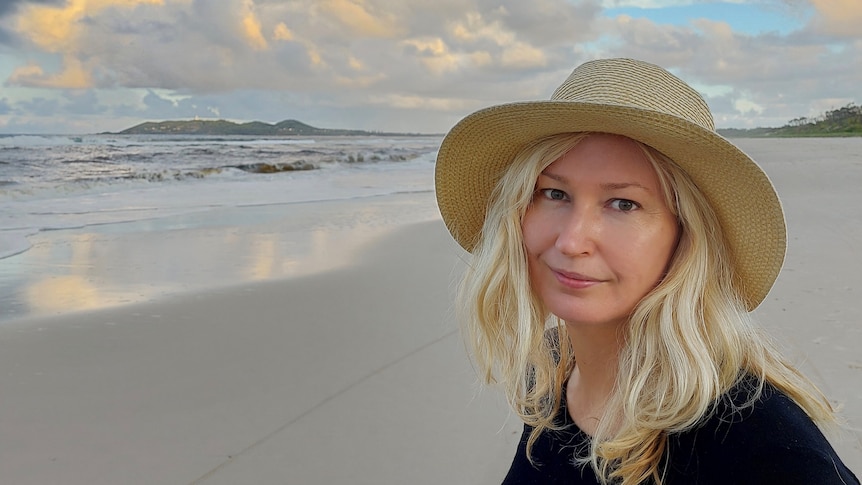 A woman in a black had with blonde hair stands on an empty beach.