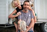 Man and woman hugging sitting in doorway of vintage caravan. Man is holding a blurred piece of concrete up to the lens.