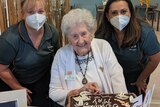 An older woman cutting a birthday cake, surrounded by two yonger women 