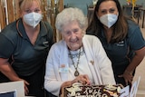 An older woman cutting a birthday cake, surrounded by two yonger women 