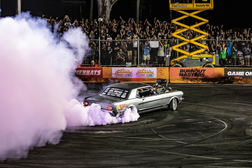 A car does a burnout while a crowd watches on from behind a safety barrier.