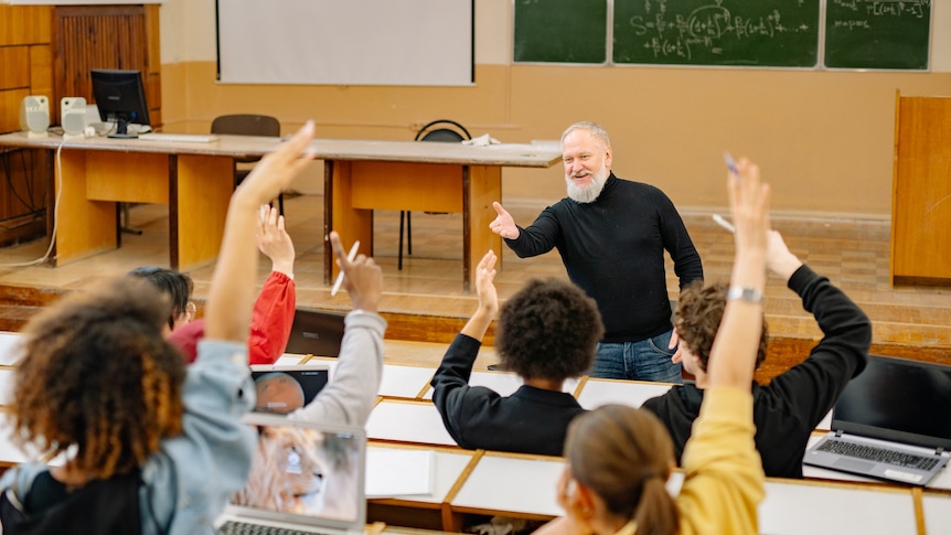 Photo of a university classroom. A group of student are facing towards the front with their hands raised with a lecture in front