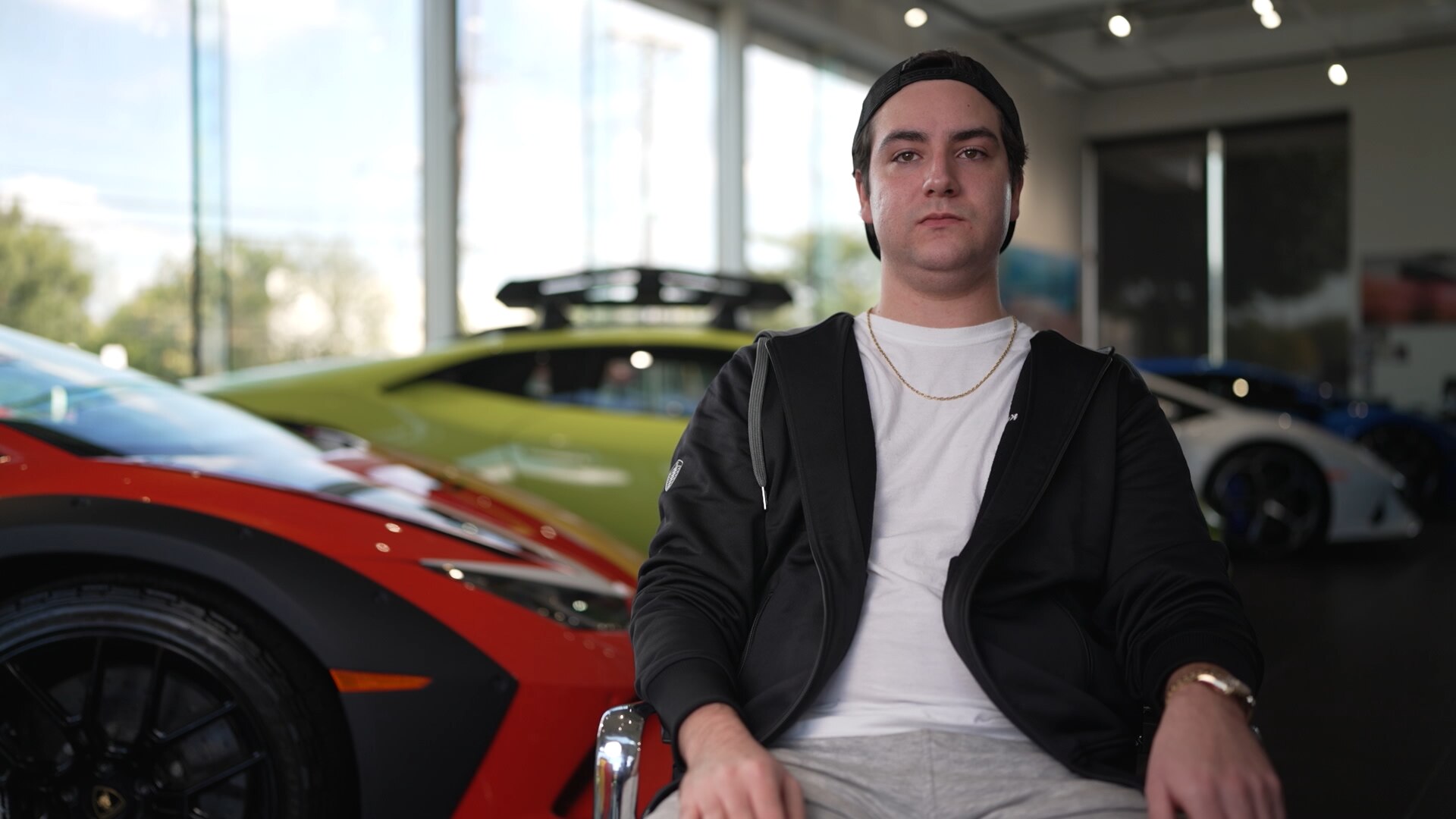 A man in his 20s wearing a white t-shirt, cap and hoodie sits in a Lamborghini dealership.