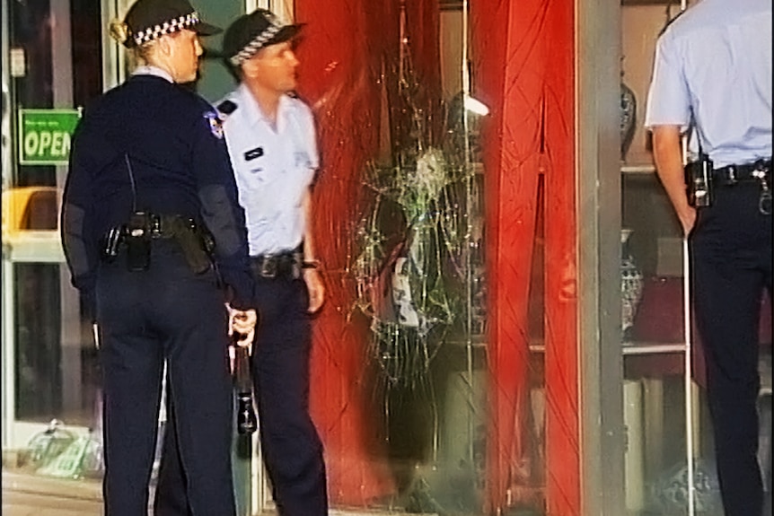 The shattered front window of a Chinese restaurant with police walking alongside.