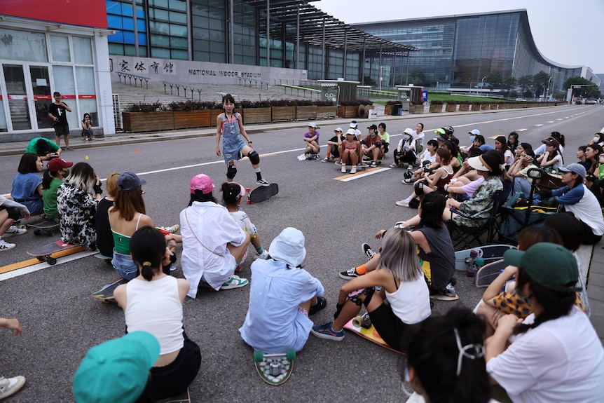 A woman stands with a sketboard adressing a group of women with skateboards who are sitting on the ground. 