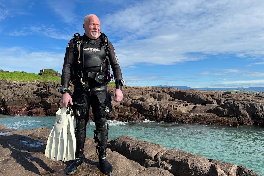 Craig stands in wetsuit and SCUBA gear on a rock shelf next to the ocean, holding flippers.