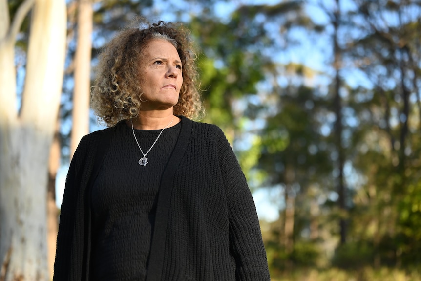 A woman wearing a black shirt stands amongst gum trees and looks off into the distance.