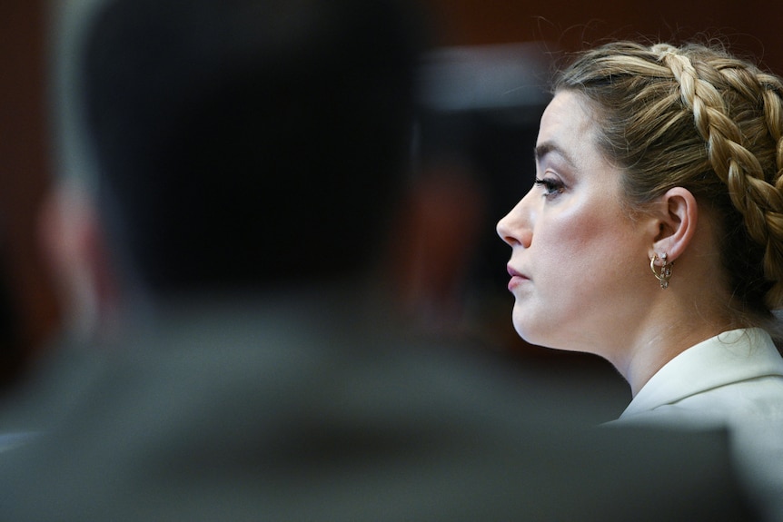 Amber Heard is seen in profile, behind an out-of-focus shadow in the foreground. She is wearing a white blazer and braid crown