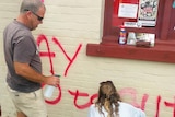 Two people scrubbing at red graffiti on the front of a building