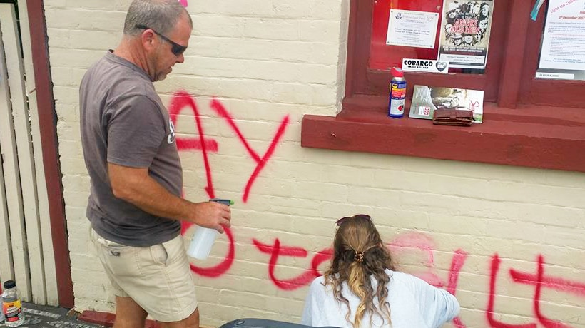 Two people scrubbing at red graffiti on the front of a building