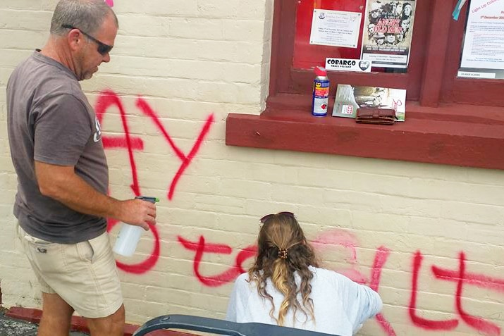 Two people scrubbing at red graffiti on the front of a building
