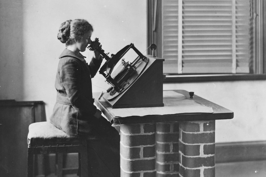 Black and white picture, woman sits up straight at a brick working bench, using an instrument to calculate the location of stars
