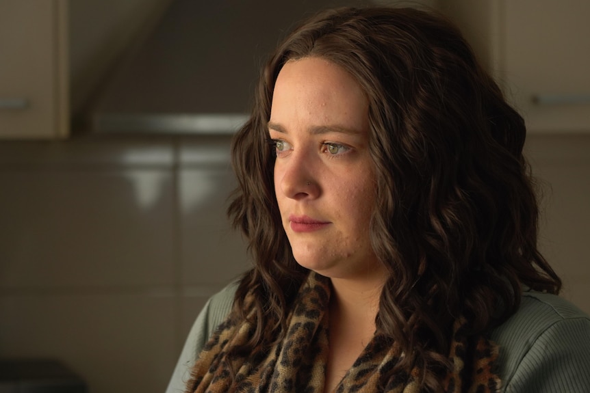 Laura, with long brown hair, stands in a kitchen looking out the window.