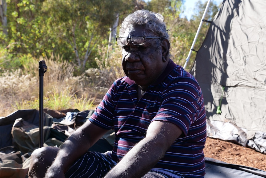 An older indigenous man sits cross legged on the ground