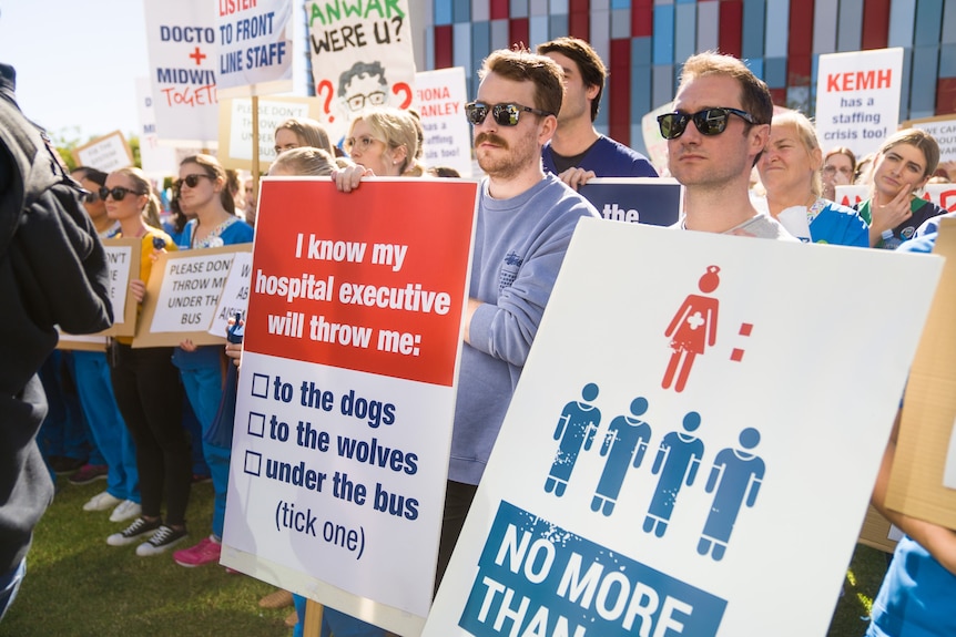A large group of people wearing medical attire, standing at a rally holding large placards and signs.