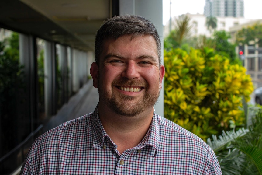 A smiling man in a checked shirt with plants behind him.