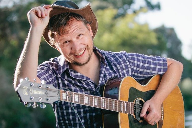 Man in check shirt, cowboy hat, lifting hat off his head slightly with cheeky grin, looking at the camera.