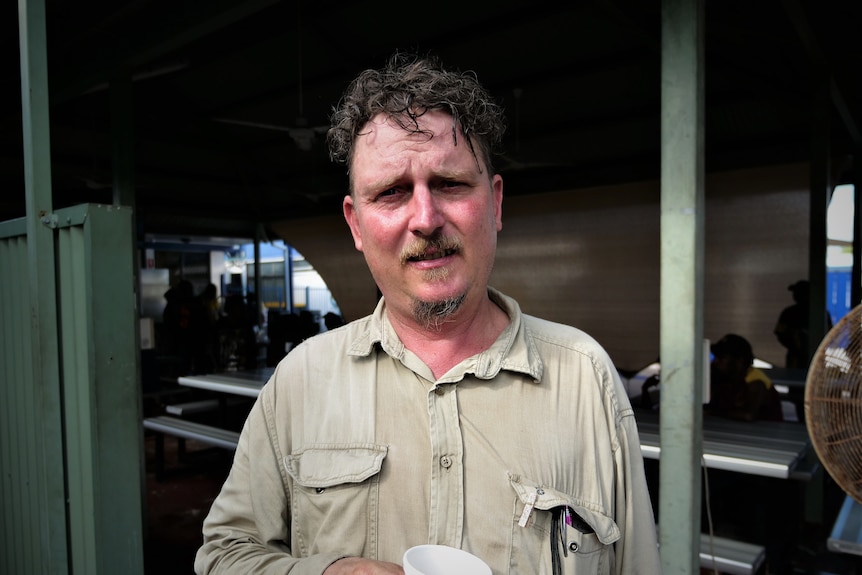 Man with curly hair standing under a shade structure. Ripped shirt. Holding polystyrene cup.