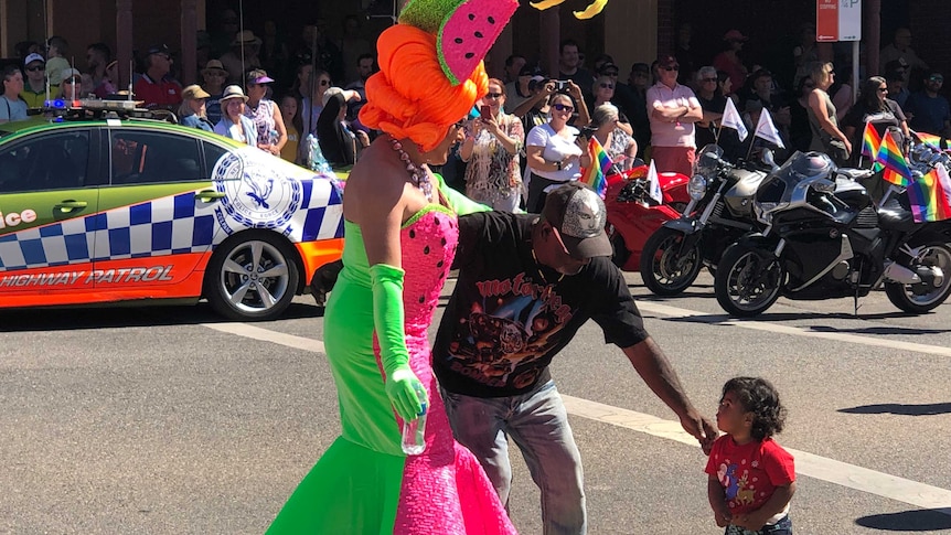 A drag queen dressed in a water melon themed costume greets a young Aboriginal child.