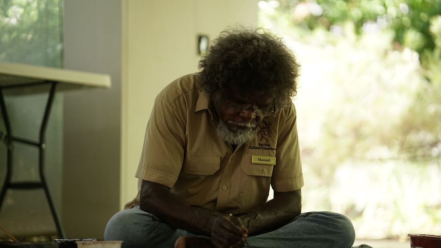 An Indigenous artist sits on the floor, wearing glasses, as he paints a picture