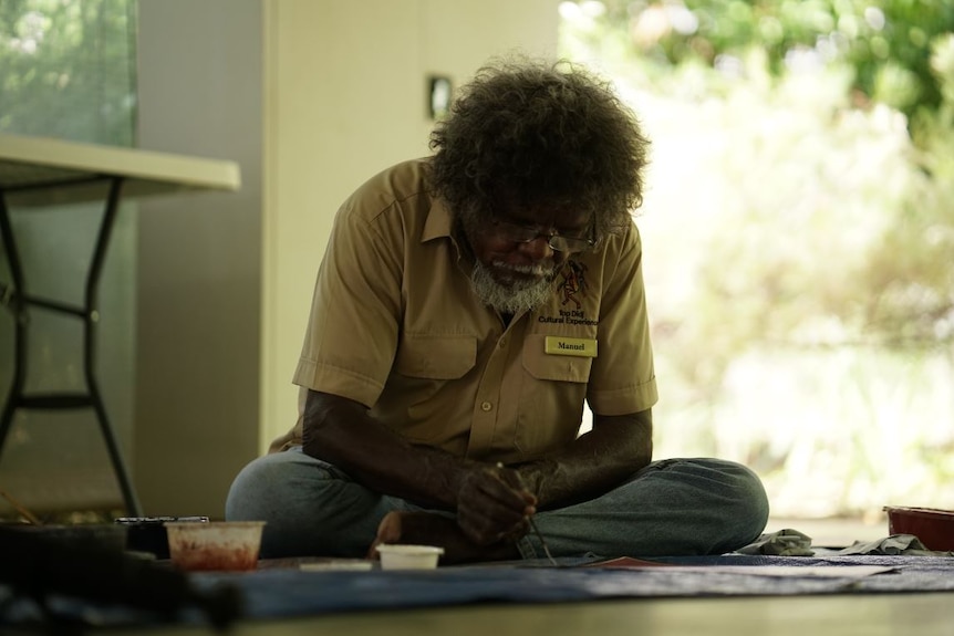 An Indigenous artist sits on the floor, wearing glasses, as he paints a picture