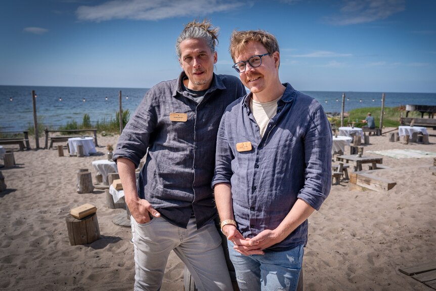 Two men in blue shirts pose for the camera near a beach on a sunny day.