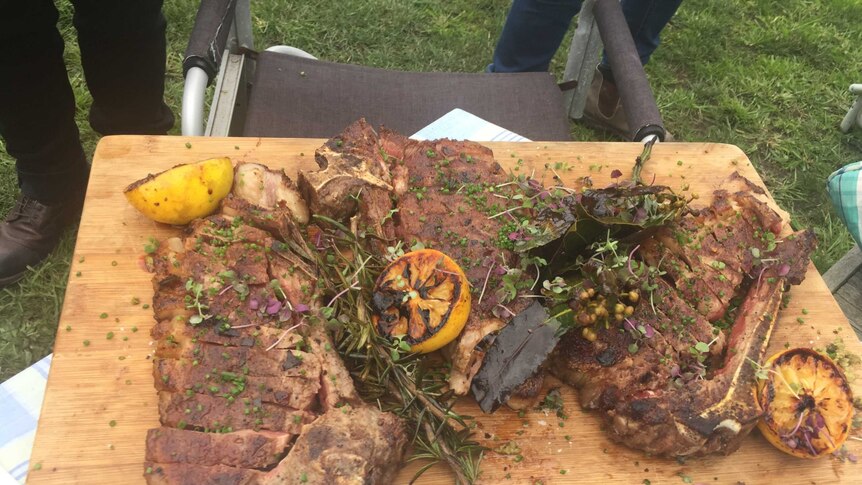 Cooked beef sitting on a wooden board with lemons and herbs scattered over it.