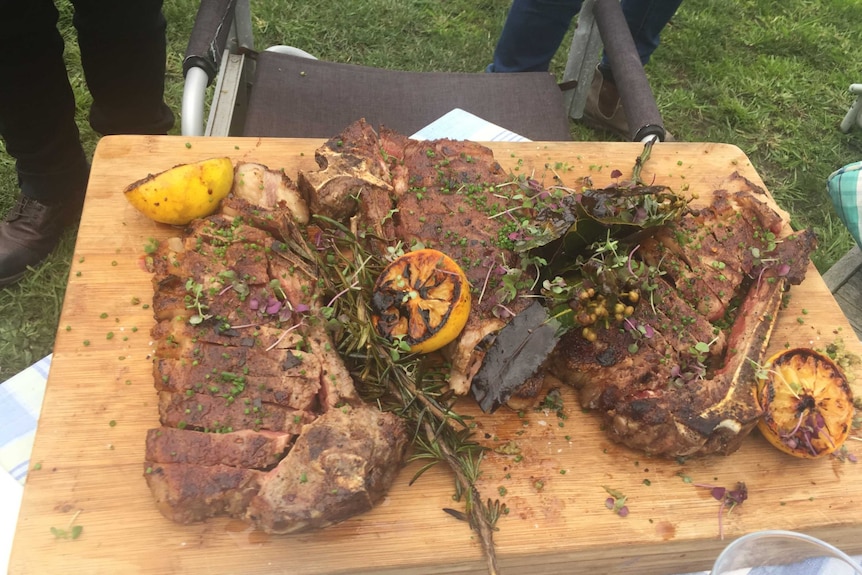 Cooked beef sitting on a wooden board with lemons and herbs scattered over it.