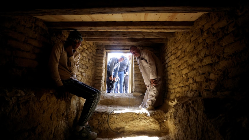 Two men sit inside a stone cave as other people look in from a small doorway. 
