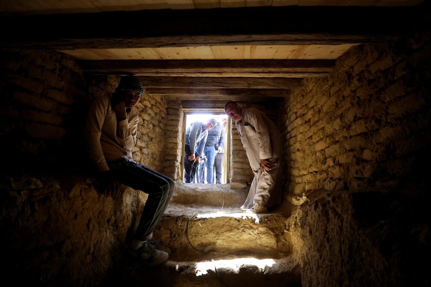 Two men sit inside a stone cave as other people look in from a small doorway. 