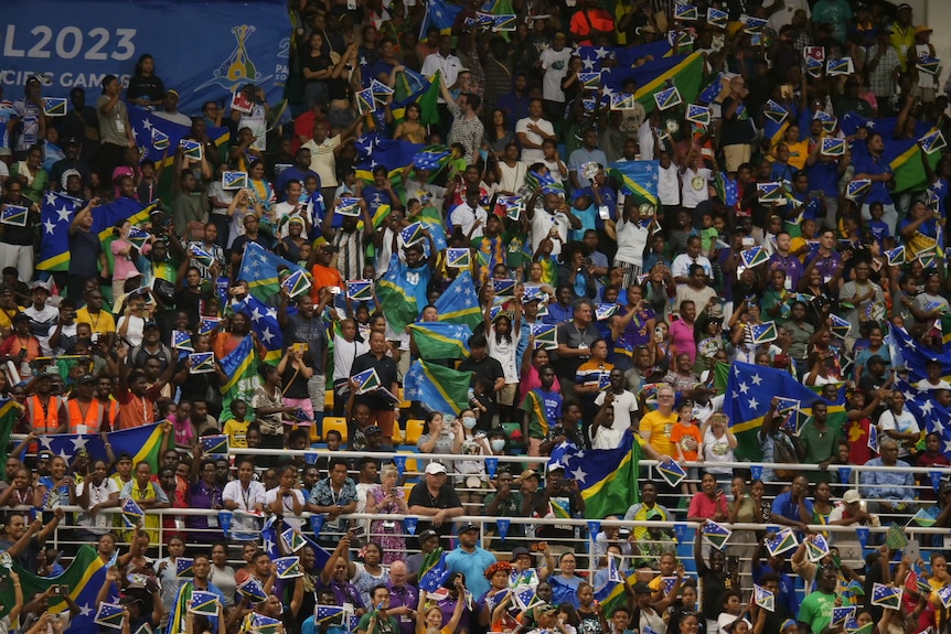 A crowd of people sitting in a stadium cheer and hold up flags.
