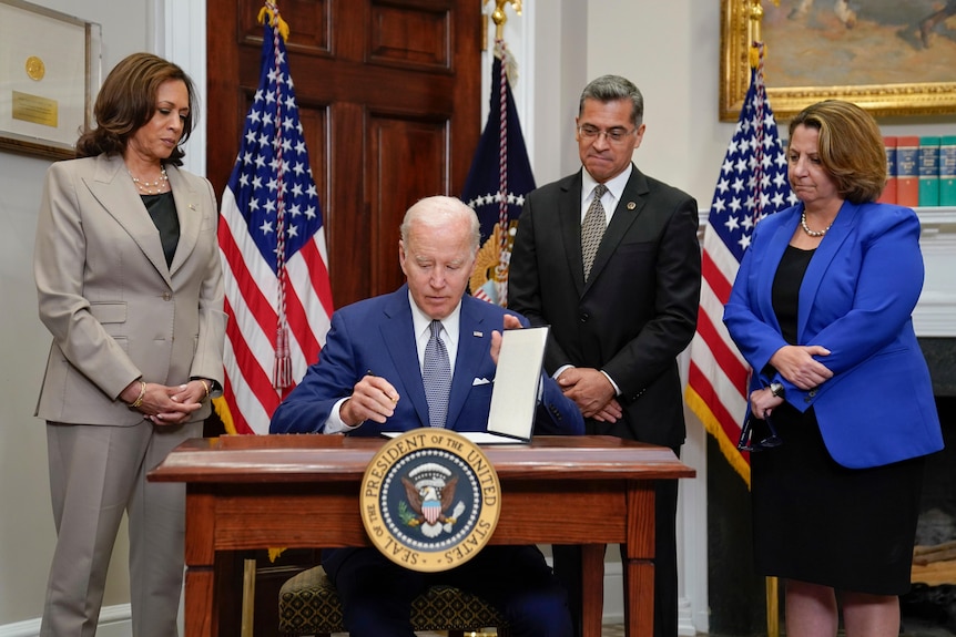 Joe Biden sitting surrounded by others including vice president Kamala Harris with American flag in the background.