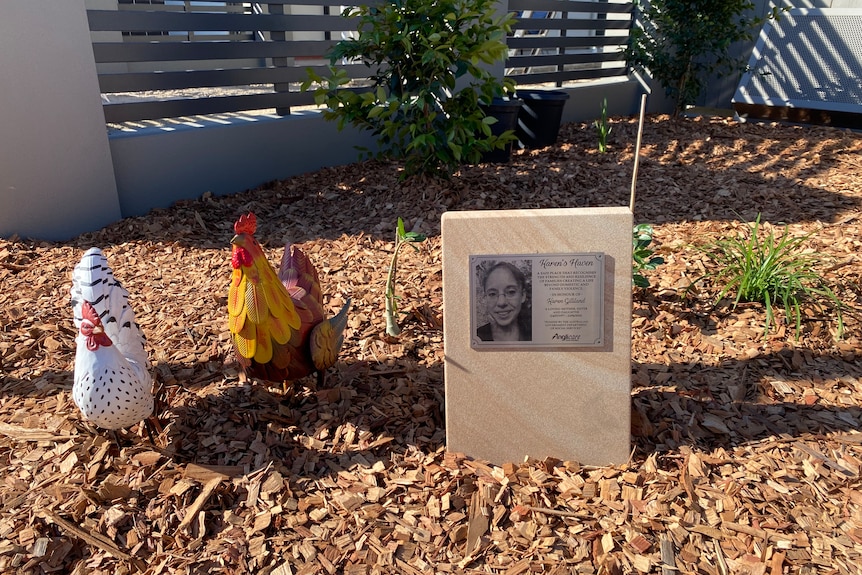 A woodchip garden bed with two chicken statues next to a plaque for Karen Gilliland.