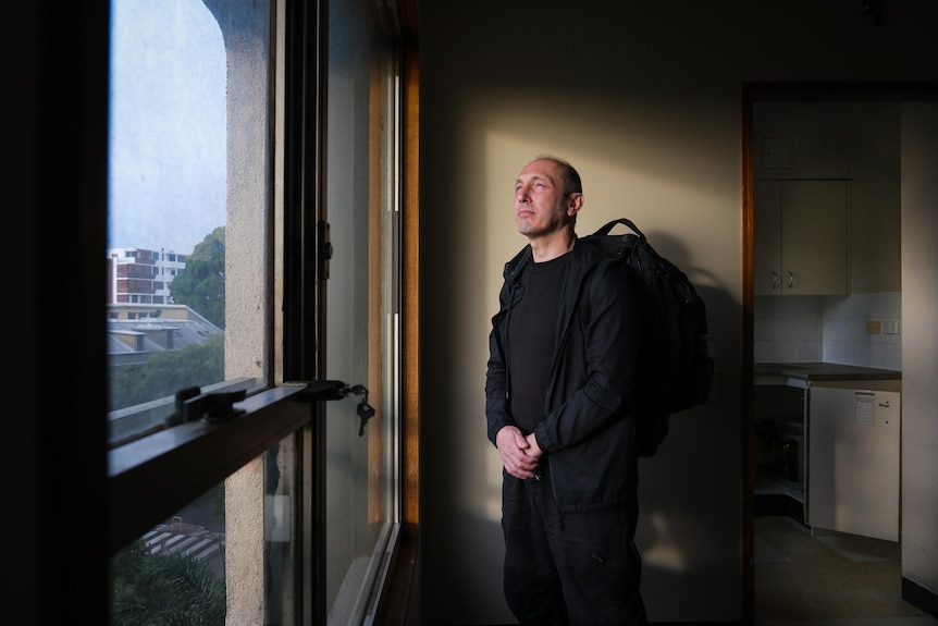 A man wearing a backpack looks out of his apartment window